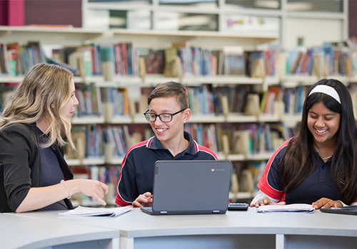 Photo of a teacher talking to 2 students sitting down in front of laptops at a library table.